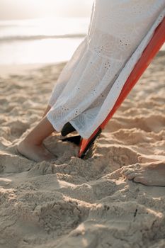 Barefoot woman in white dress stands on sandy beach with surfboard.