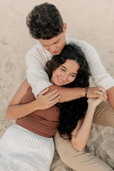 A happy couple sitting and embracing each other on a sandy beach, enjoying a sunny day.
