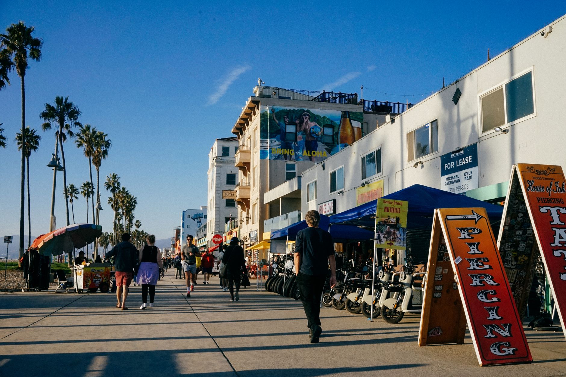 a busy Southern California boardwalk with people strolling past small shops, ocean in the distance
