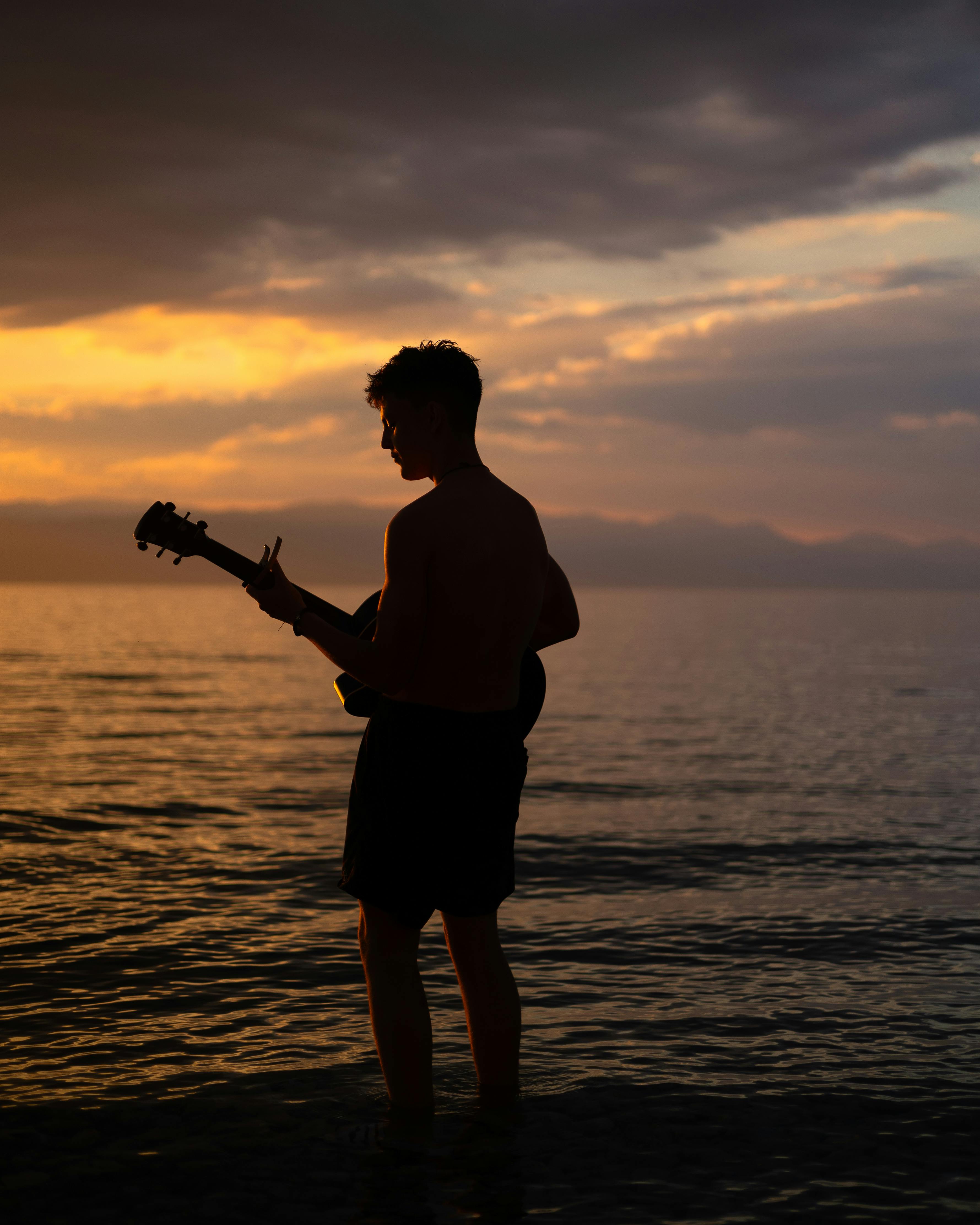 A serene silhouette of a guitarist standing in shallow water at sunset, creating a peaceful mood.