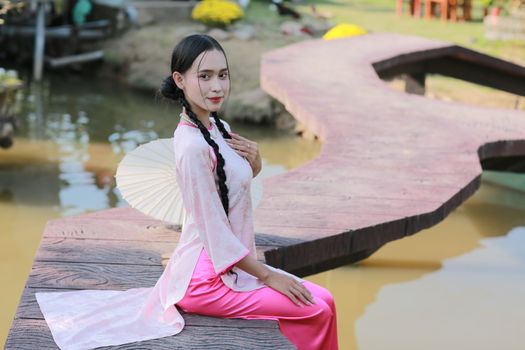 Woman in traditional attire sits by a serene pond with a parasol, exuding elegance.