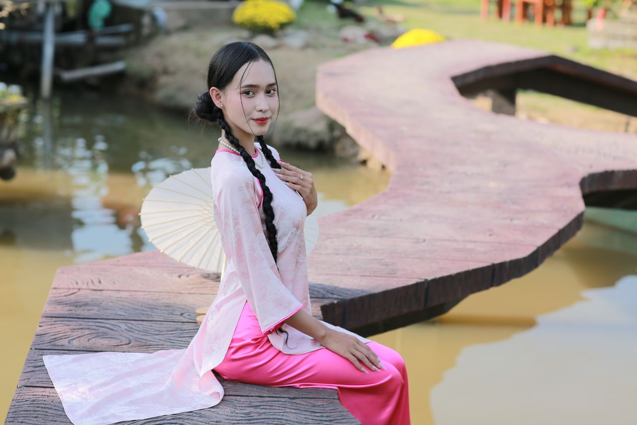 woman in traditional attire sits by a serene pond with a parasol exudi