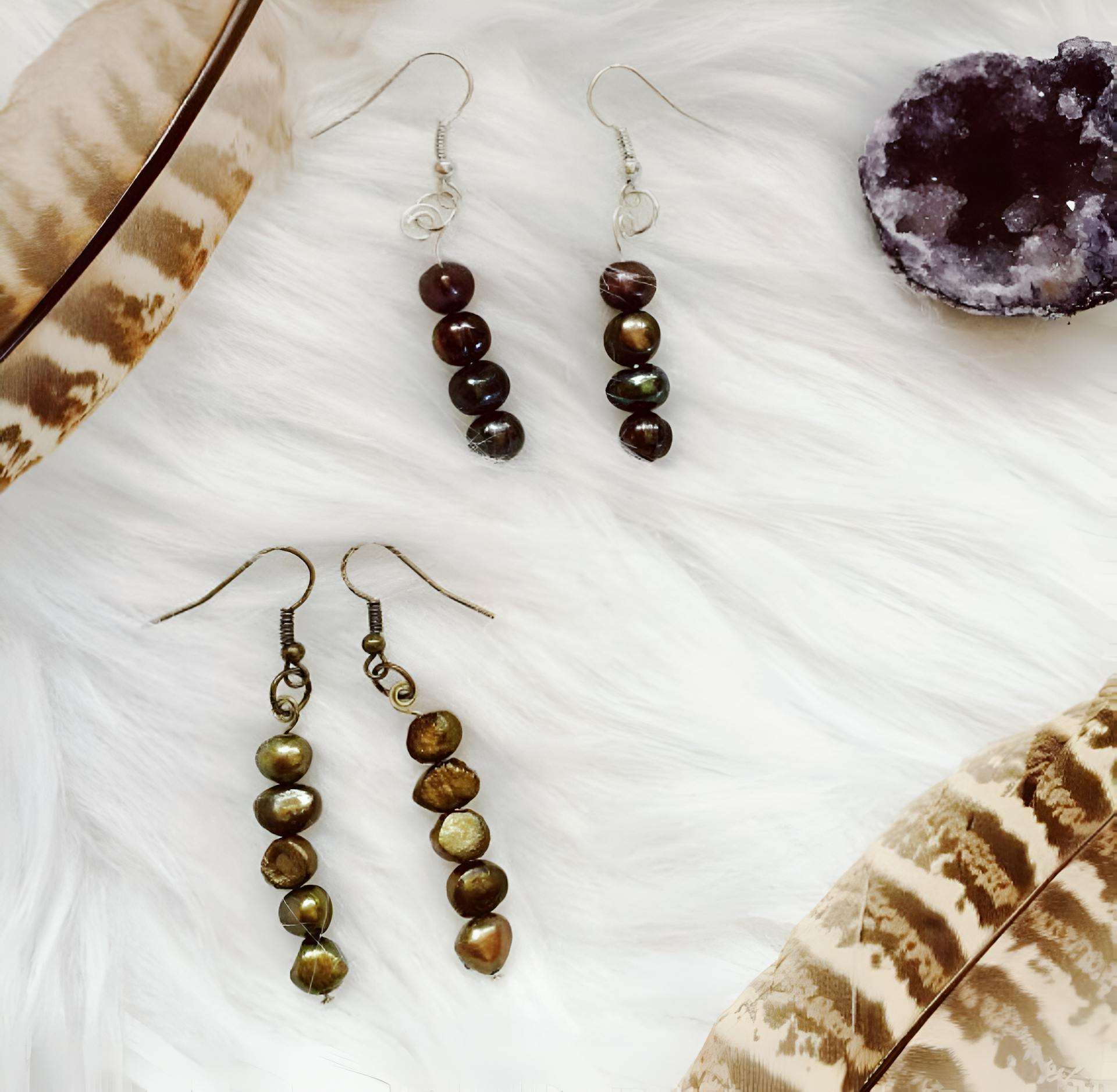 Elegant beaded earrings displayed on soft white fur beside feathers and a geode crystal.