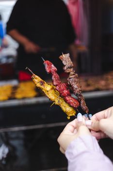 Three flavorful grilled meat skewers held in a hand at a busy street market, showcasing a variety of spices and cooking styles.