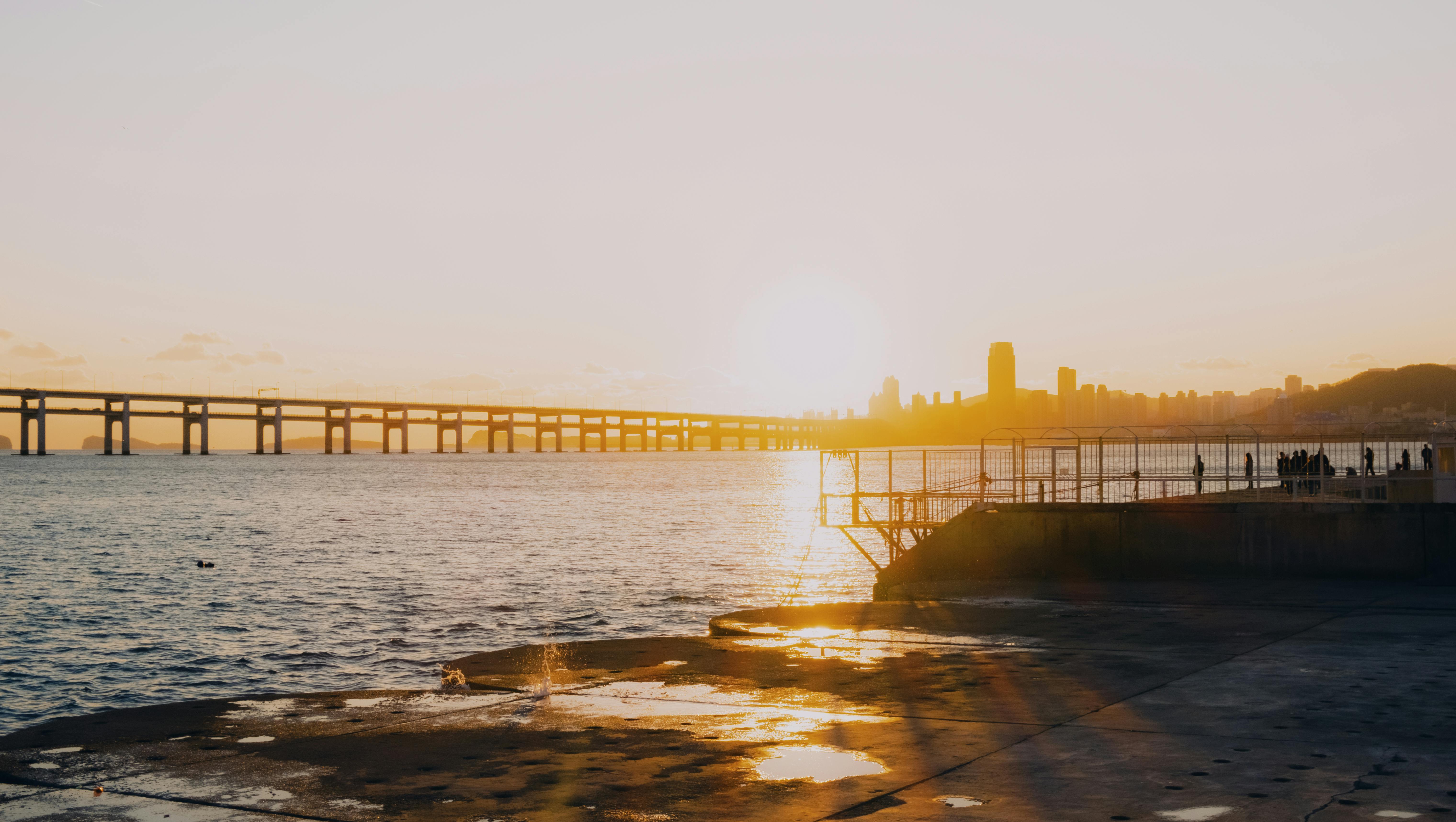 Serene sunset scene with ocean, bridge, cityscape, and silhouetted people, evoking tranquility and b