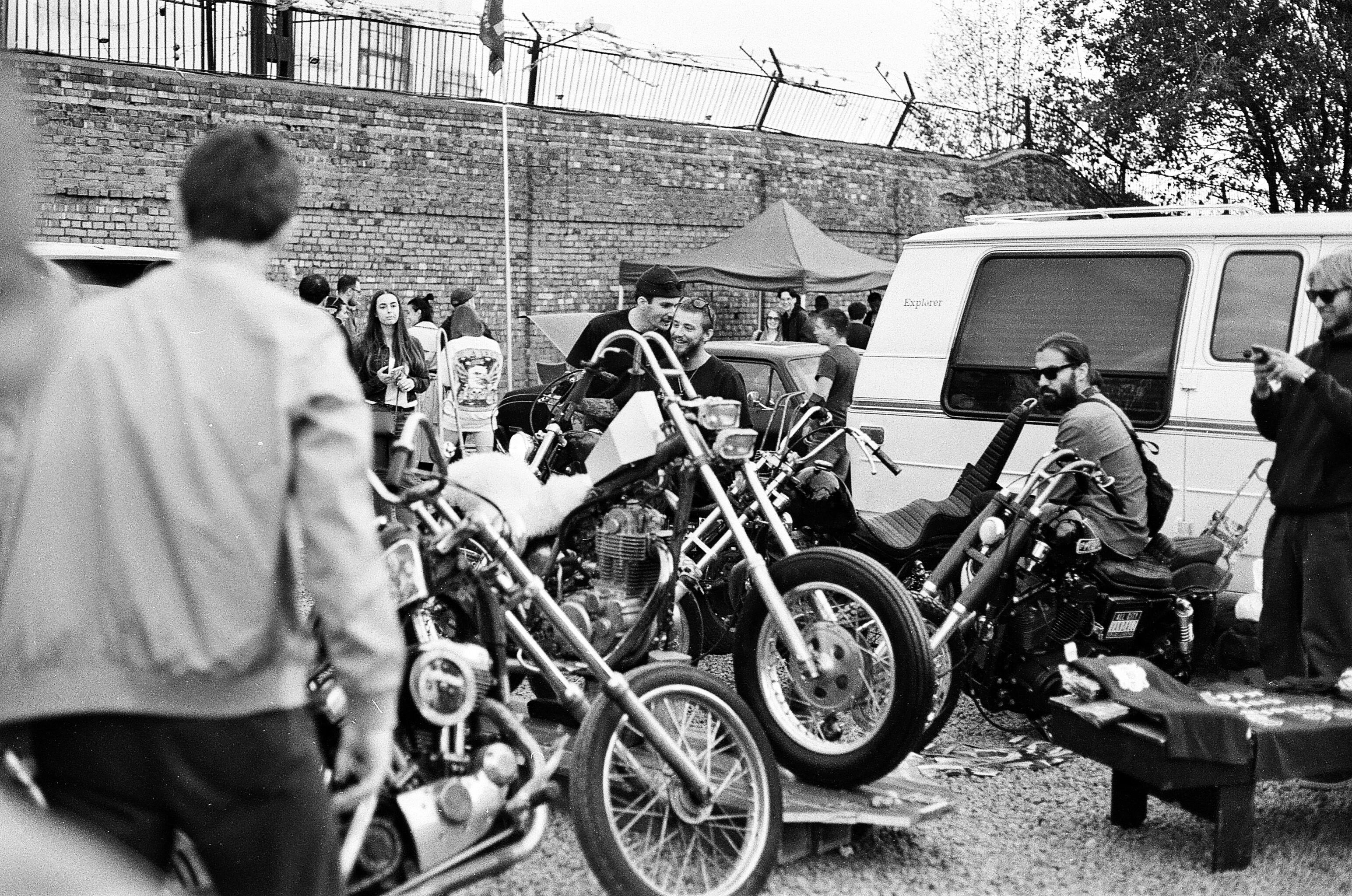 A black and white photo of a vintage biker gathering featuring multiple motorcycles and people outdoors.