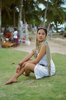 A woman in white dress sits on grass by a tropical beach, enjoying a sunny day.
