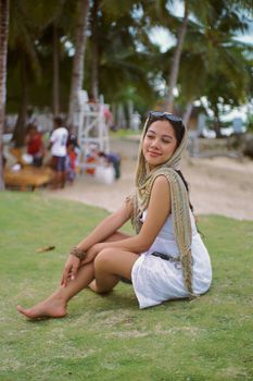 Young woman sitting on grass at a tropical beach, enjoying a sunny day.