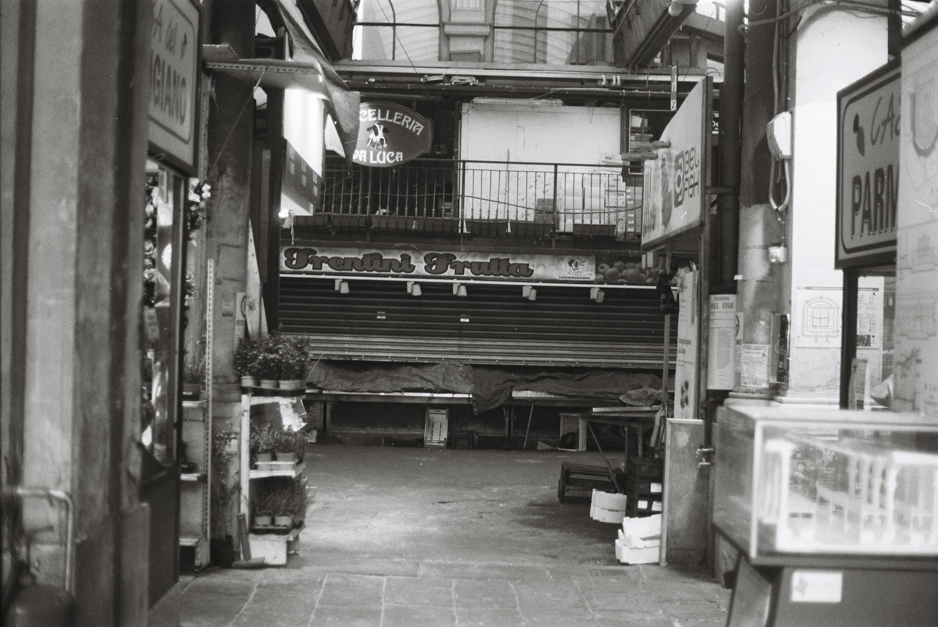 Black and white image of an alley with closed market stalls, Italian signage.