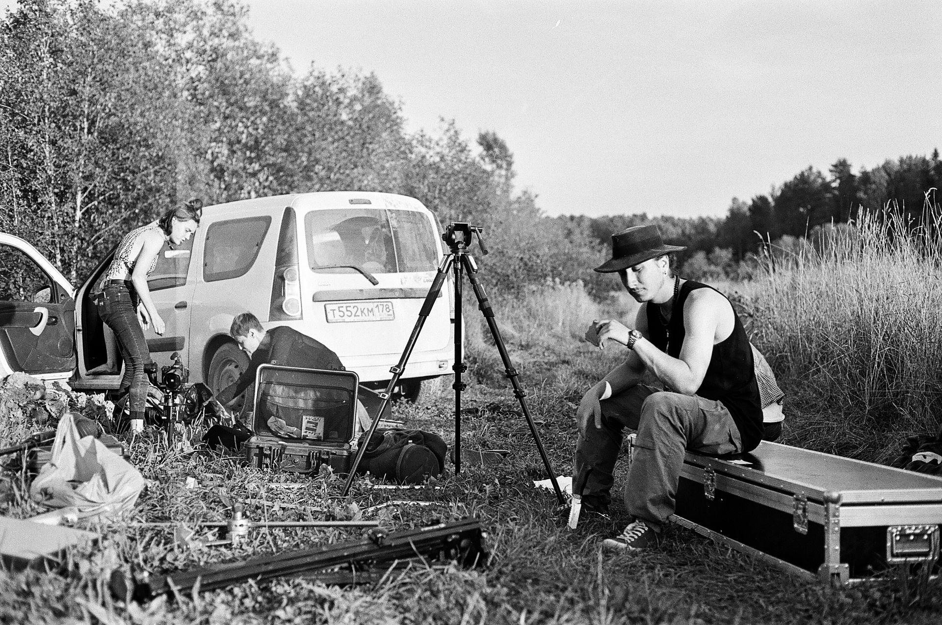 A black and white image of a film crew setting up equipment outdoors with a car and tripod.