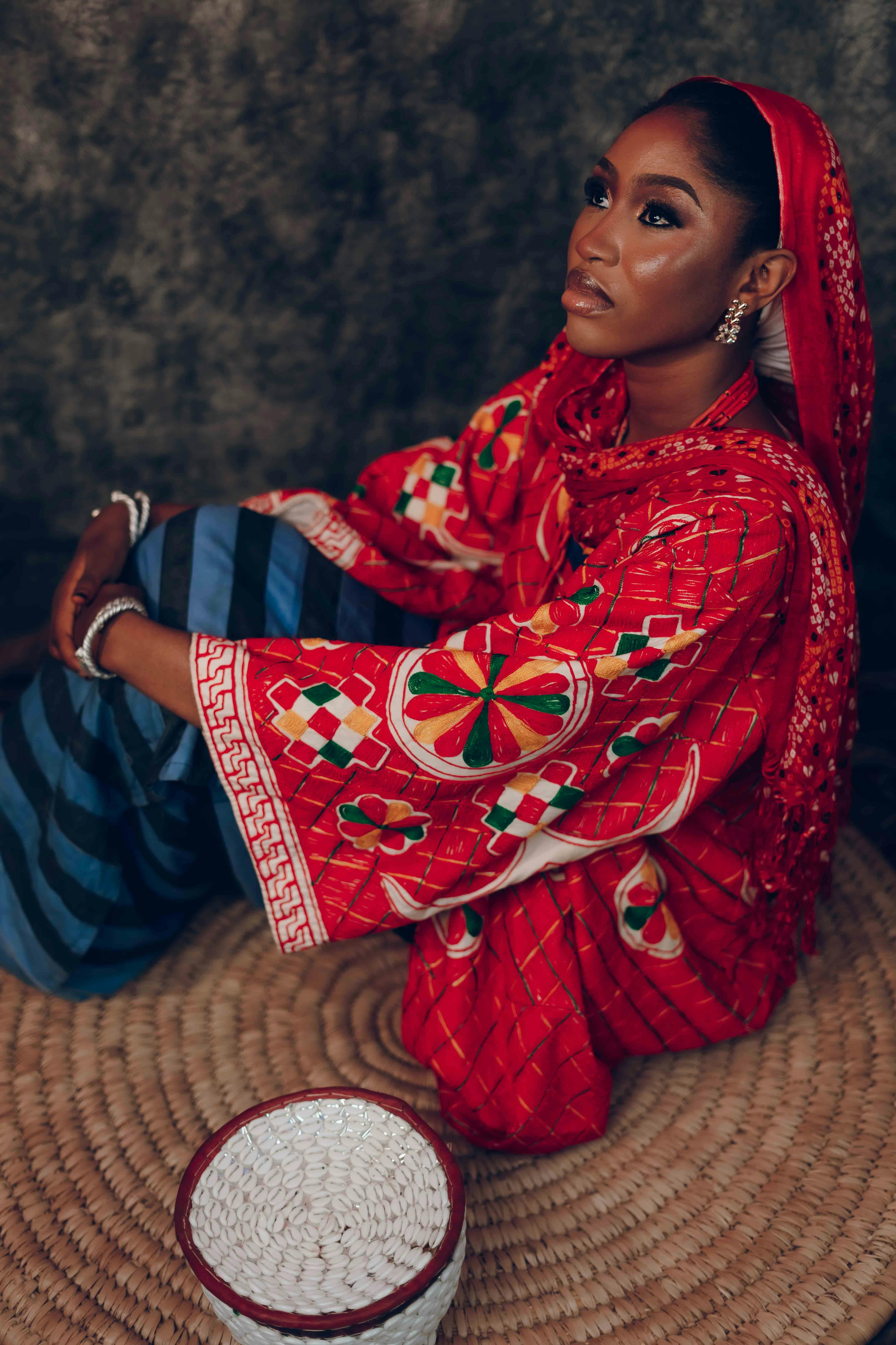 A woman in vibrant traditional African clothing sits thoughtfully indoors.