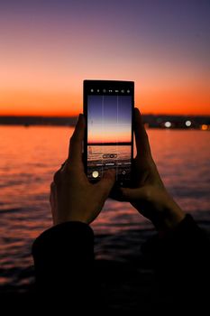 Hands holding a smartphone capturing a vivid sunset over the ocean.