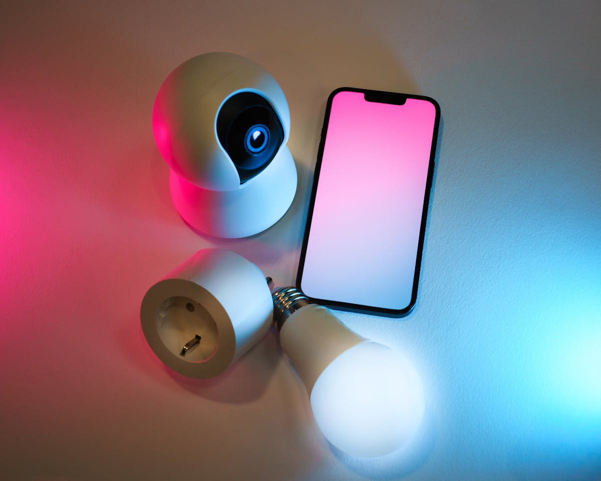Smartphone affiancato a lampadina smart, telecamera e presa domotica sul tavolo