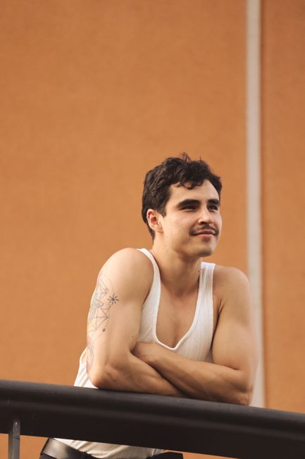 A man in a white tank top leans on a balcony railing outdoors in a relaxed pose.