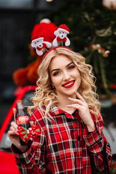 Cheerful woman celebrating holidays, holding a festive drink, wearing a red plaid shirt and holiday headband.