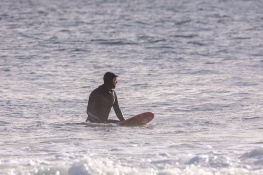 A lone surfer in a wetsuit waits patiently on his surfboard amid calm ocean waters.
