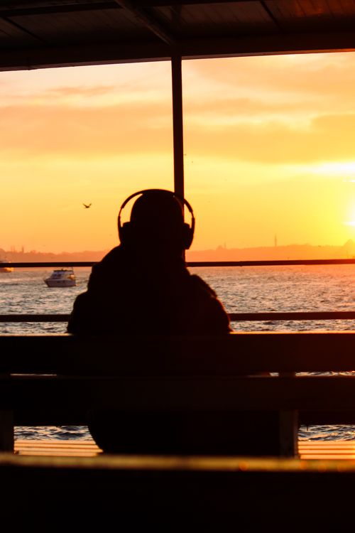 Free A person with headphones watches a vibrant sunset over the water. Stock Photo