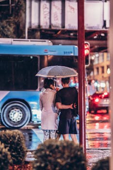 A couple stands under an umbrella, embracing on a rainy urban street at night.