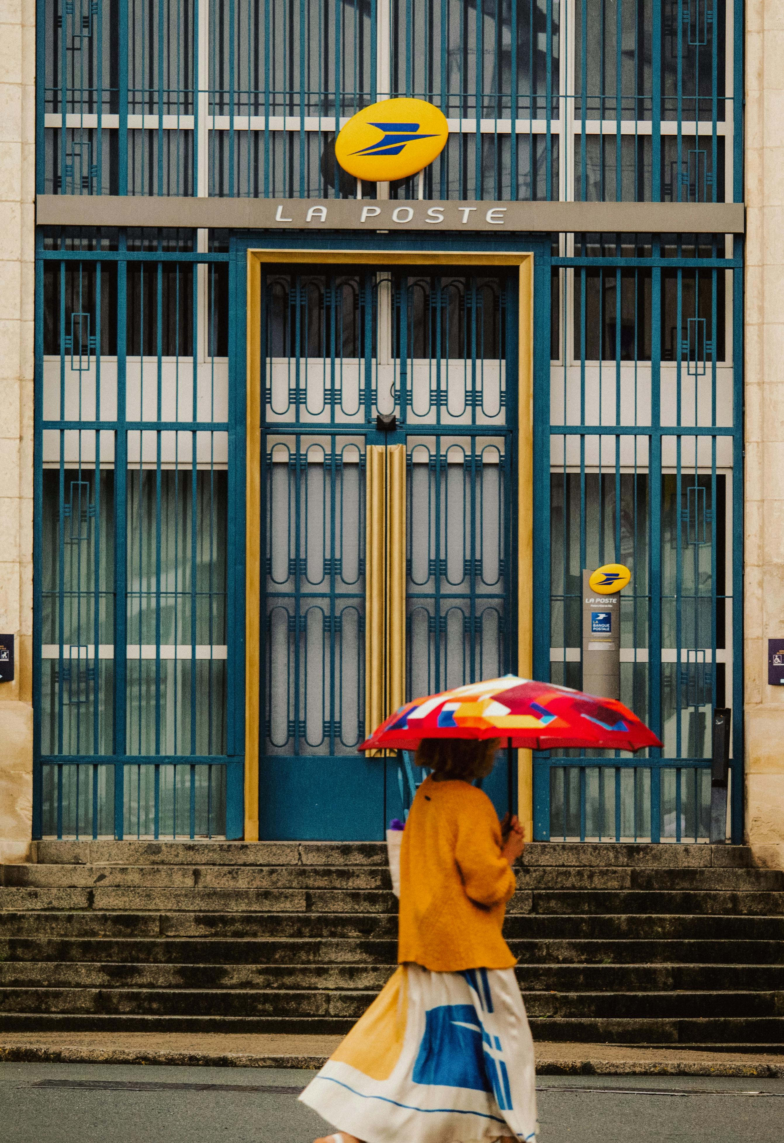 Person with a colorful umbrella walking past a French post office.