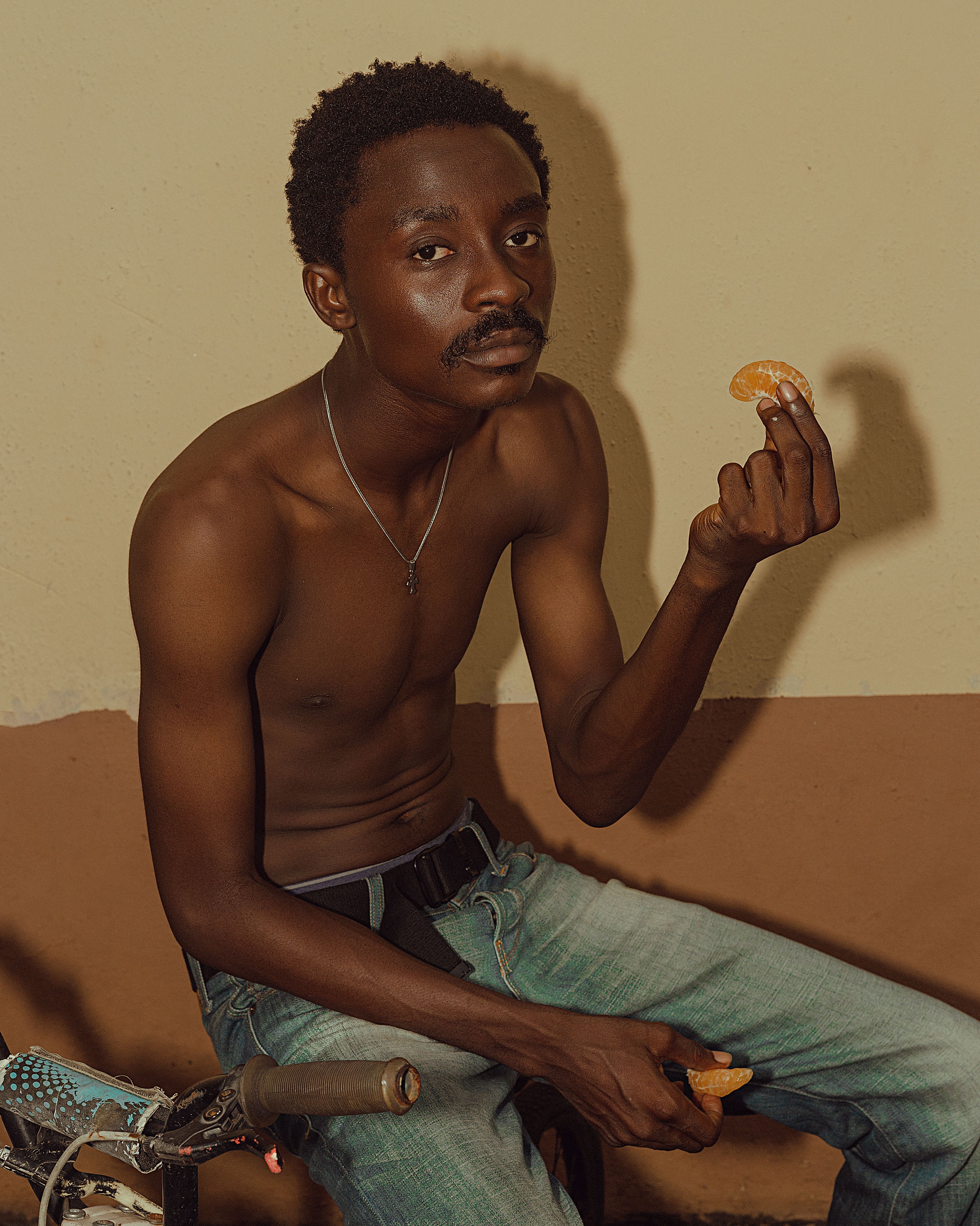 Shirtless young man sitting indoors enjoying a snack.