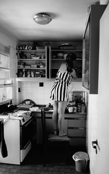 A woman stands on a chair in a kitchen, reaching for items on a high shelf.
