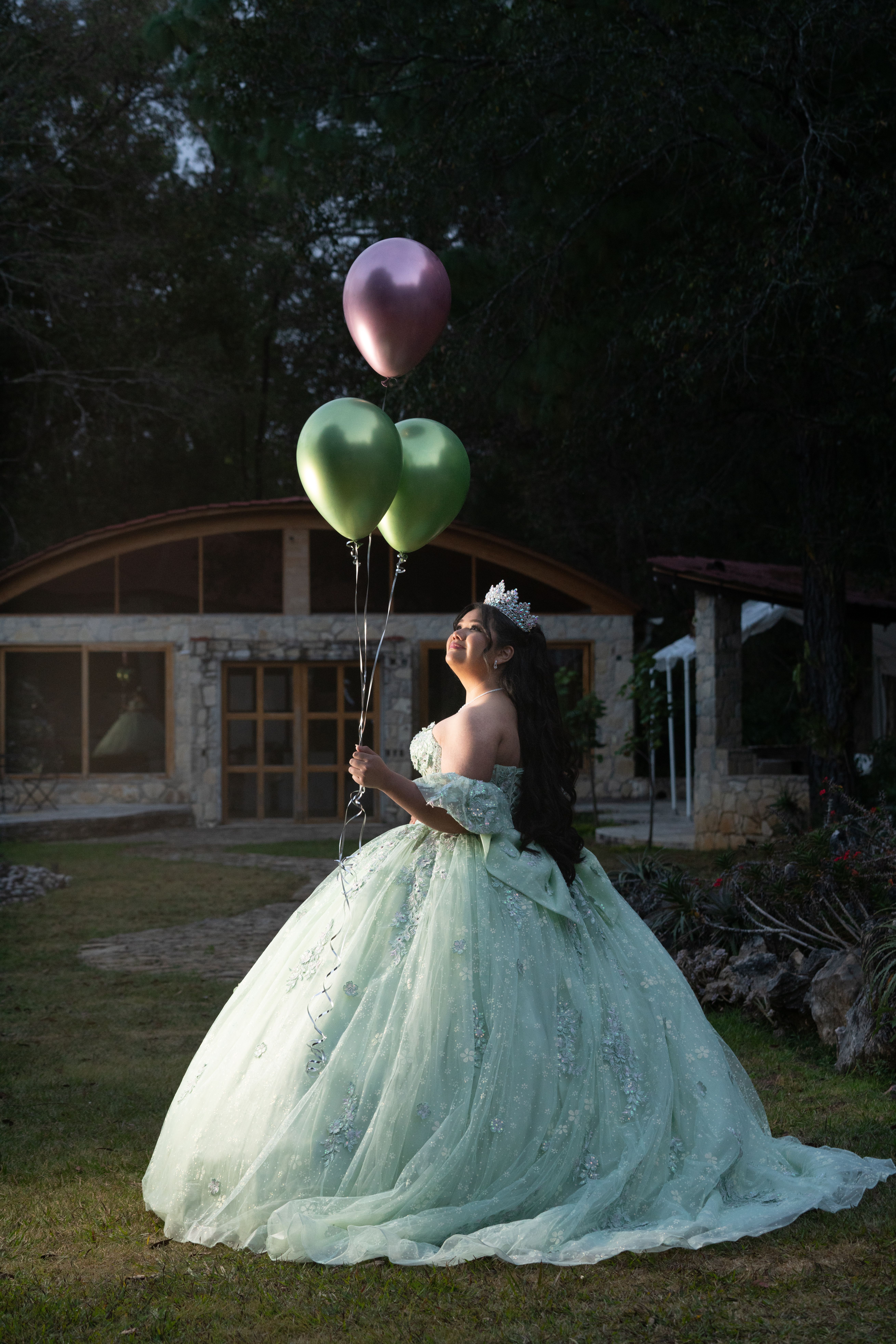 A young woman in a ball gown holds vibrant balloons in a serene garden setting.