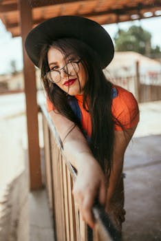 一位戴着太阳帽和眼镜的时尚年轻女子，在户外靠着栏杆摆姿势，脸上挂着自信的傻笑