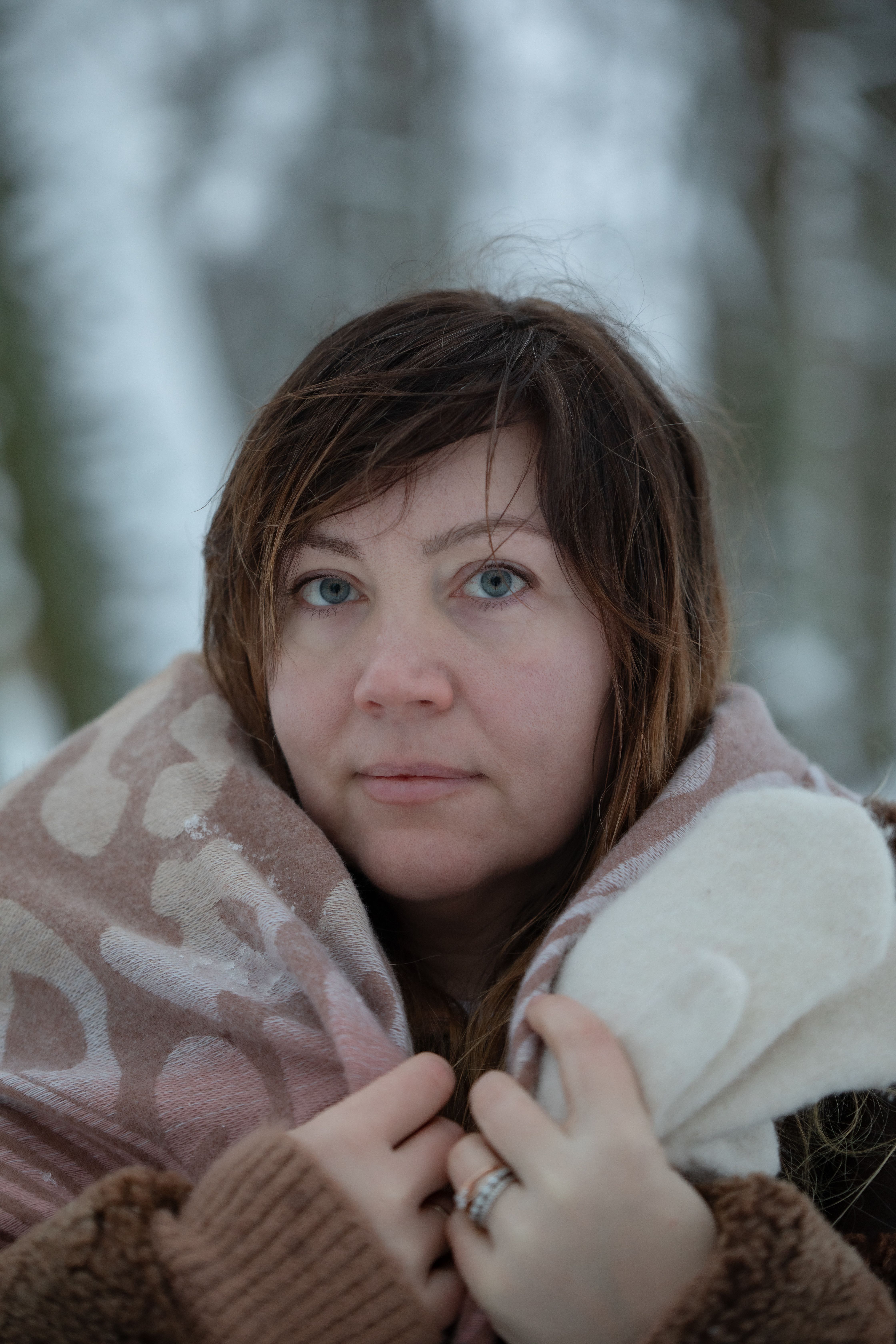 A woman with light eyes wrapped in a warm scarf, standing in a snowy forest.