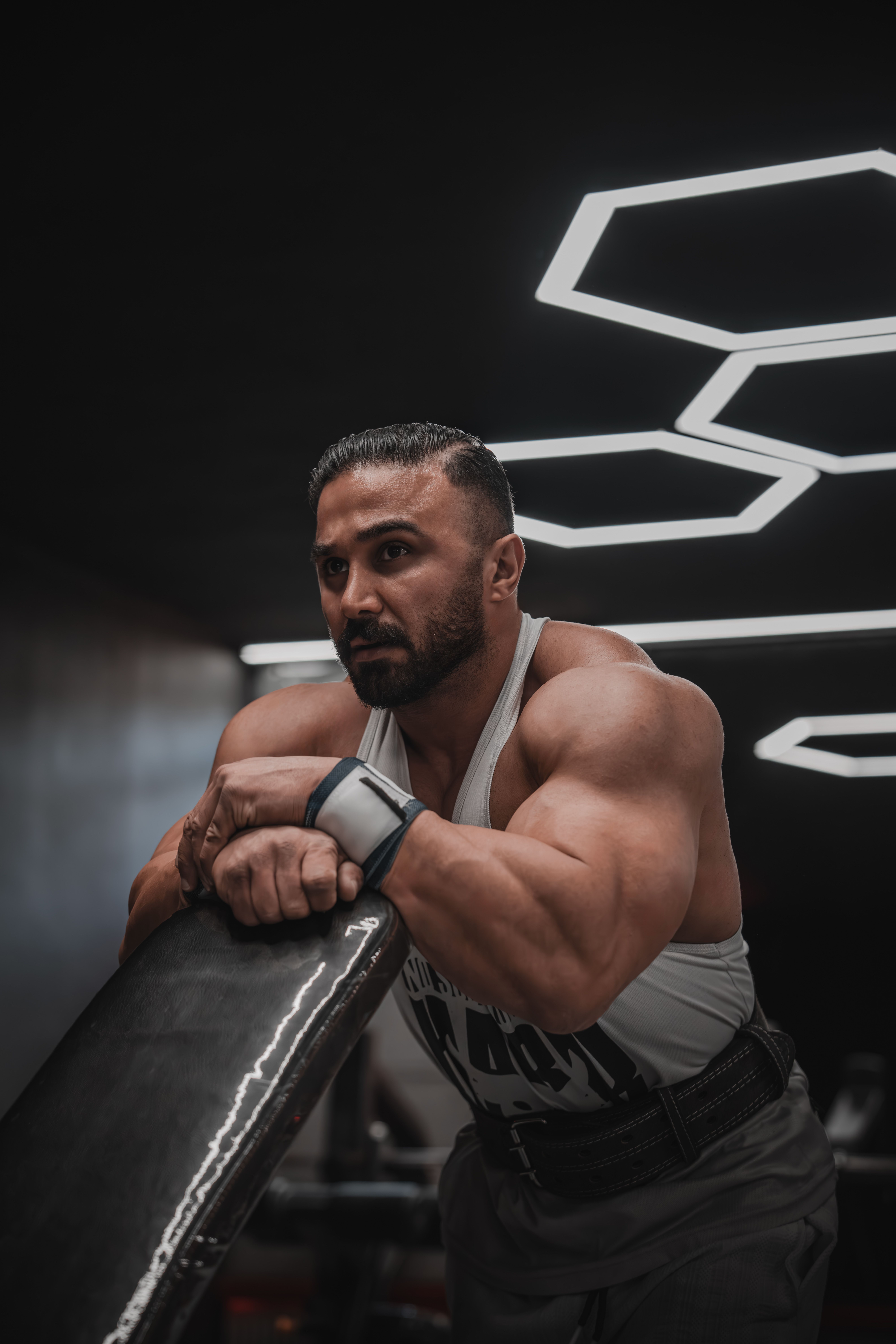 A muscular man resting in a dimly lit modern gym with geometric lights overhead.