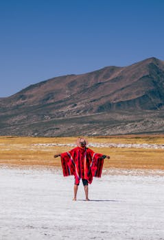 Man in a red poncho and hat stands with open arms before Peru's Andes salt flats.