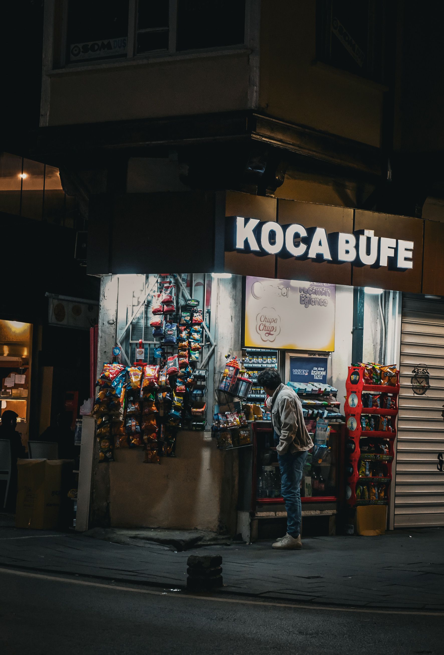 A person browsing at Koca Büfe, a corner shop, illuminated at night.