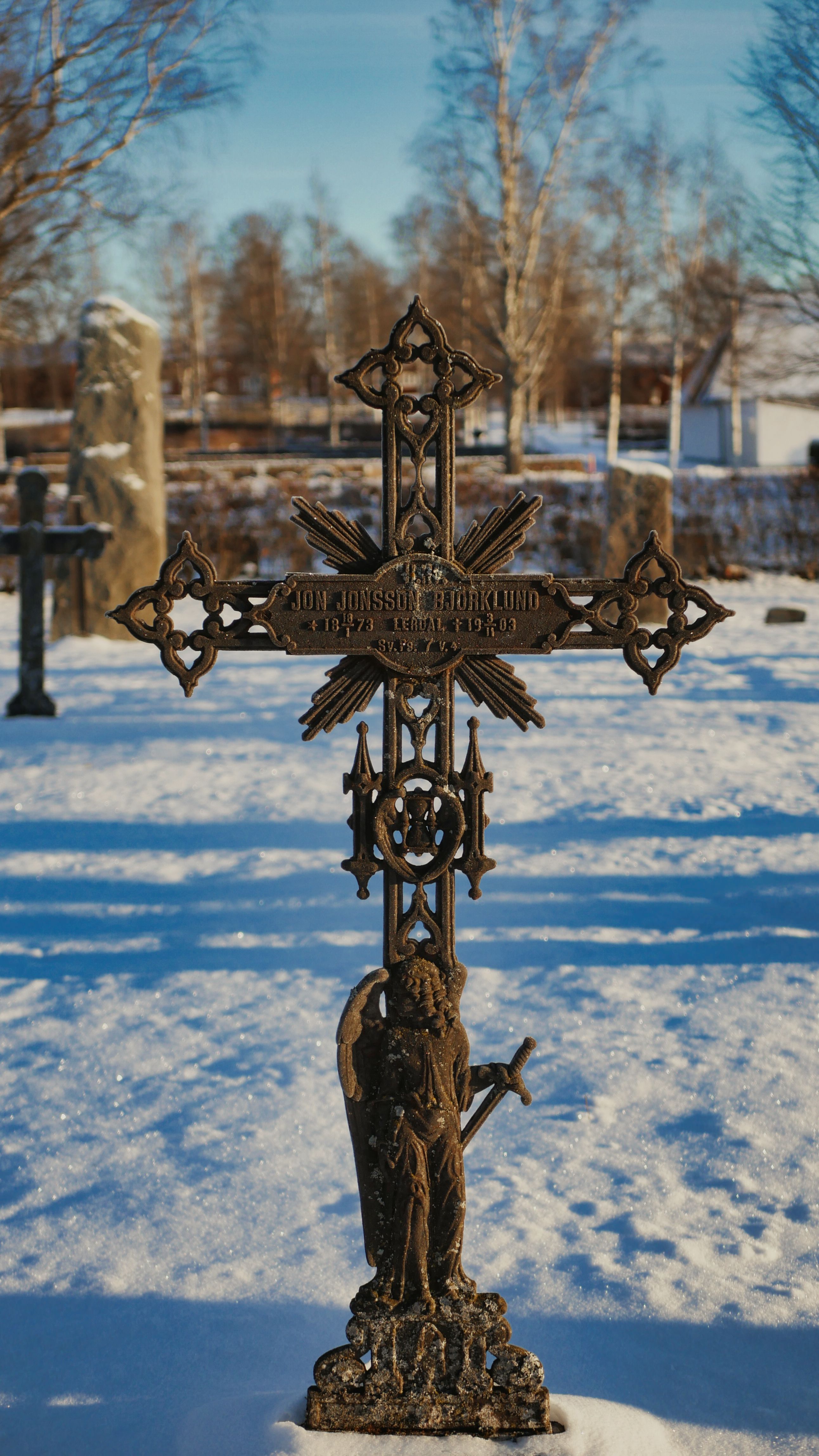 A detailed iron cross stands in a snowy Rättvik graveyard under clear winter skies.