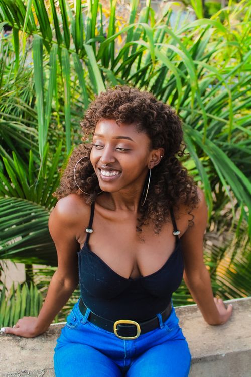 Free Fashionable woman with curly hair sitting outdoors, surrounded by vibrant plants, exuding joy and style. Stock Photo