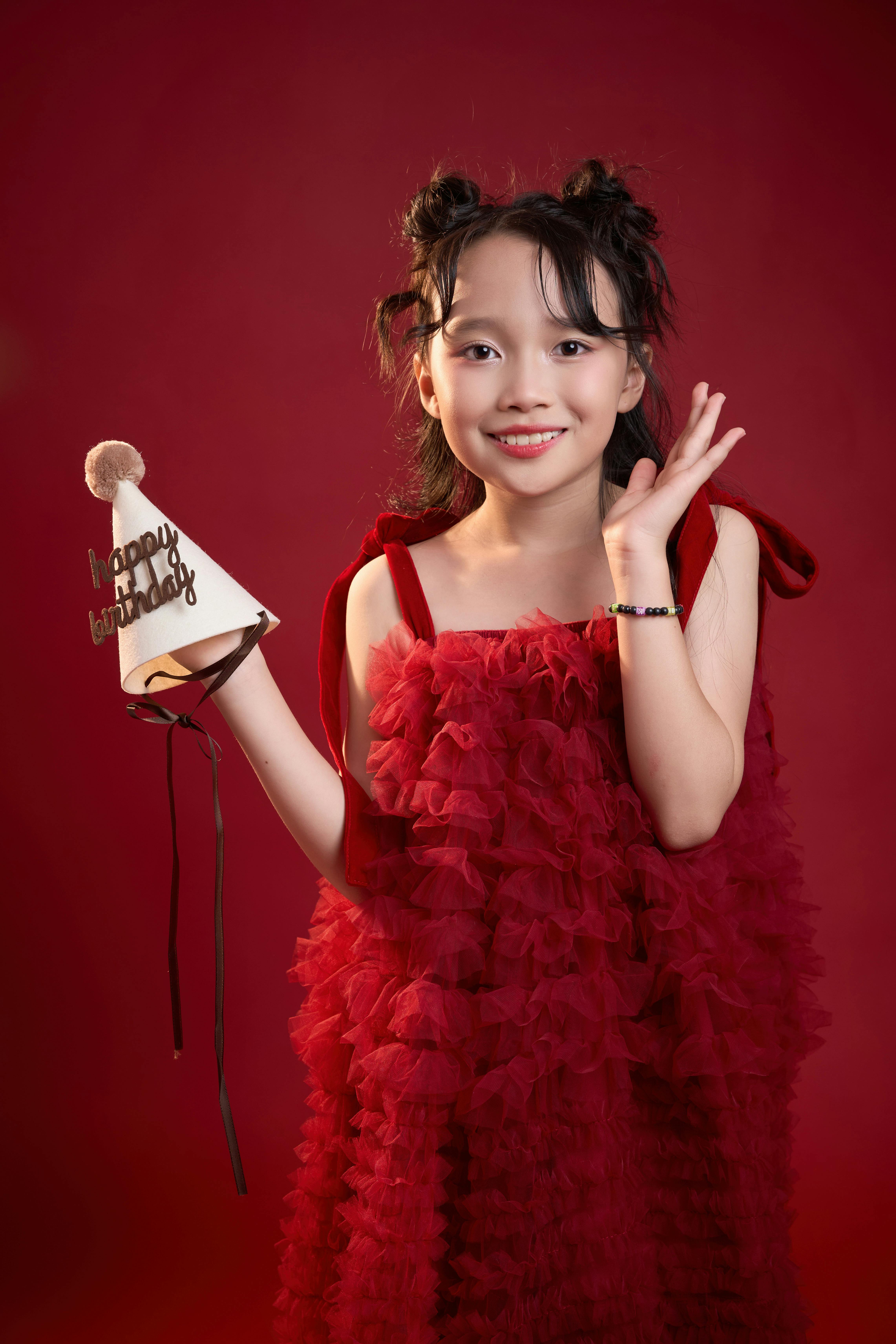 Cheerful girl in a red dress holding a birthday hat on a red background.