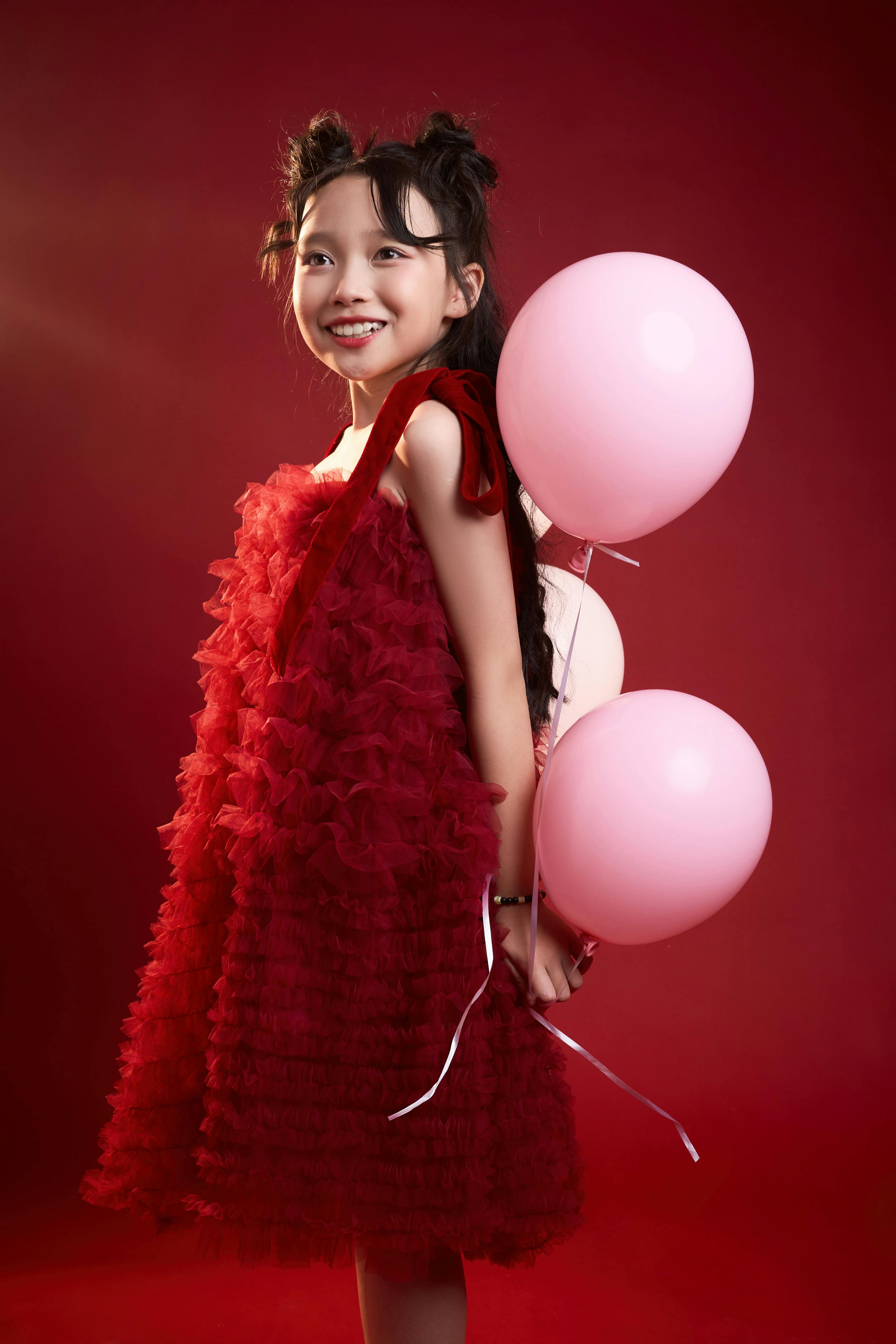 grátis Uma garota alegre em um vestido vermelho vibrante segurando balões rosas sobre um fundo vermelho. Foto profissional
