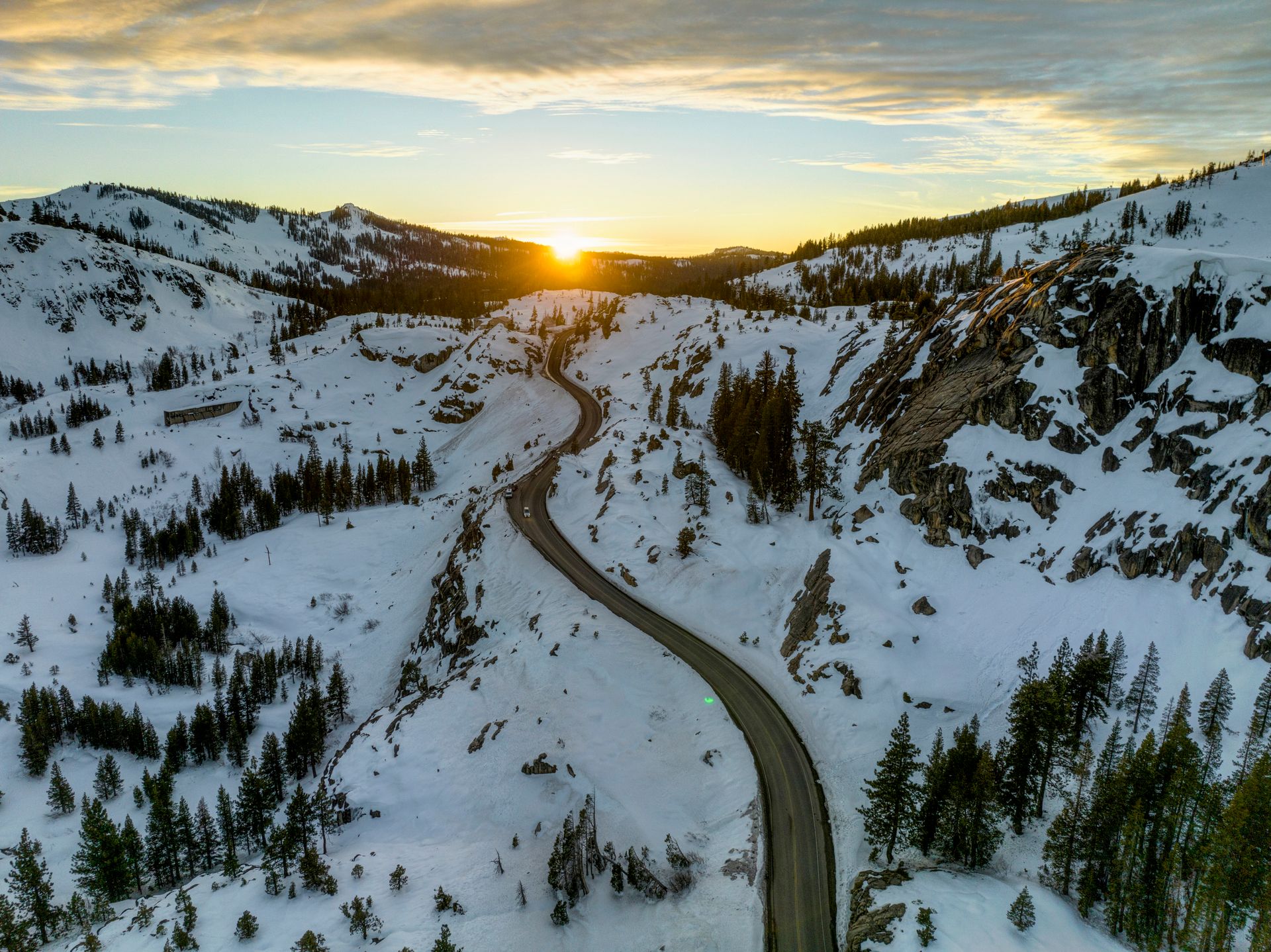 A mesmerizing aerial view of a snowy Sierra mountains landscape at sunset, showcasing a winding road amidst winter wilderness.