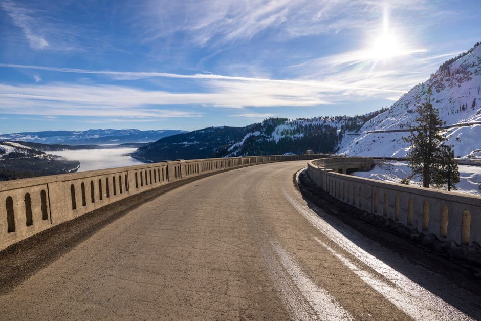 Snowy road curving through Donner Pass with a sunny sky and mountainous backdrop.
