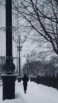 Person jogging on a snowy city sidewalk with street lamps and bare trees. Winter scene.