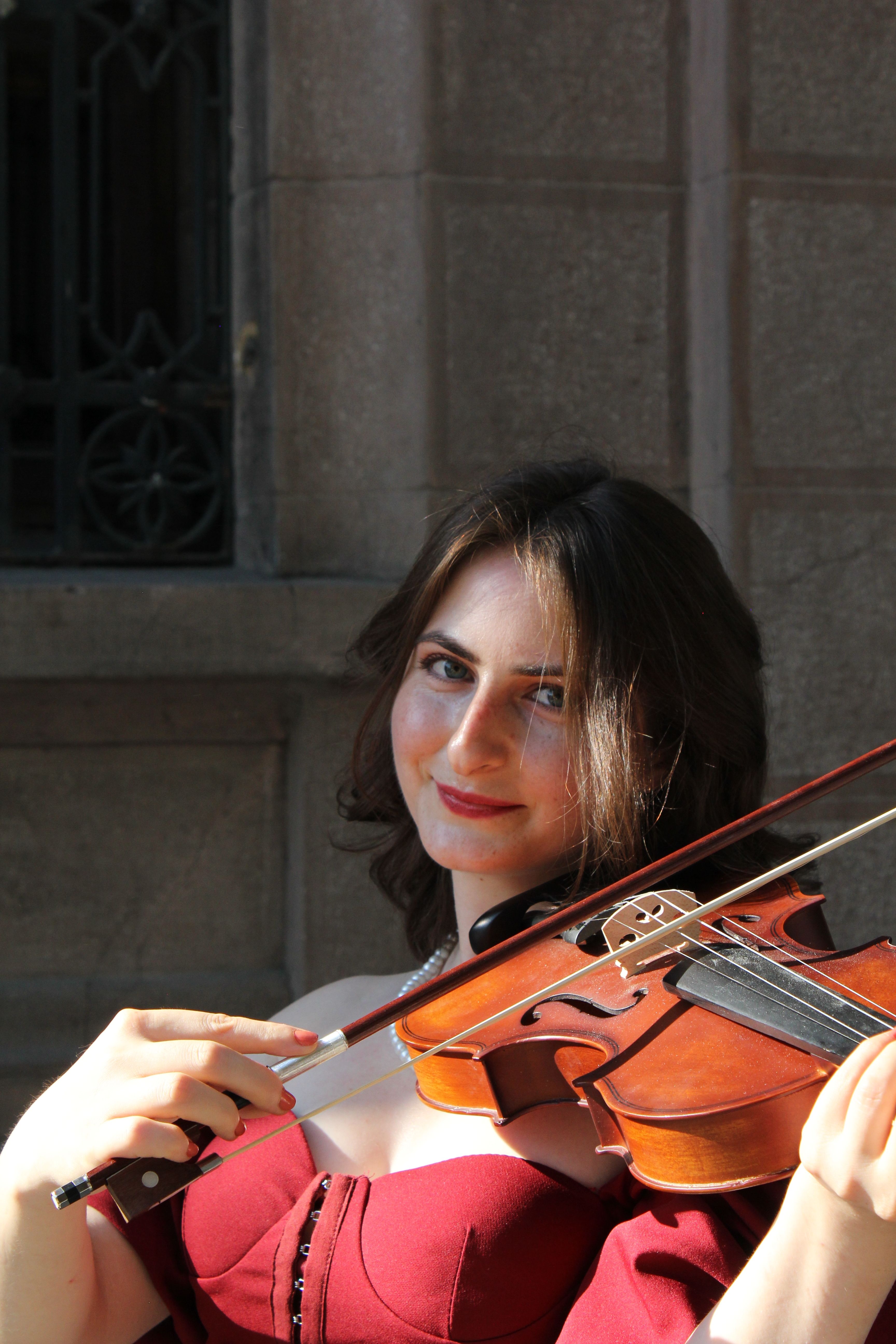 A woman in a burgundy dress plays a violin outside, smiling warmly in the sunlight.