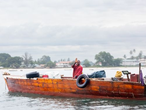 Free A fisherman waves from a wooden boat on a cloudy day with a scenic coastal background. Stock Photo