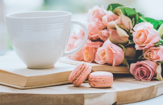 A charming coffee setup with pink roses, macarons, and an open book on a white backdrop.
