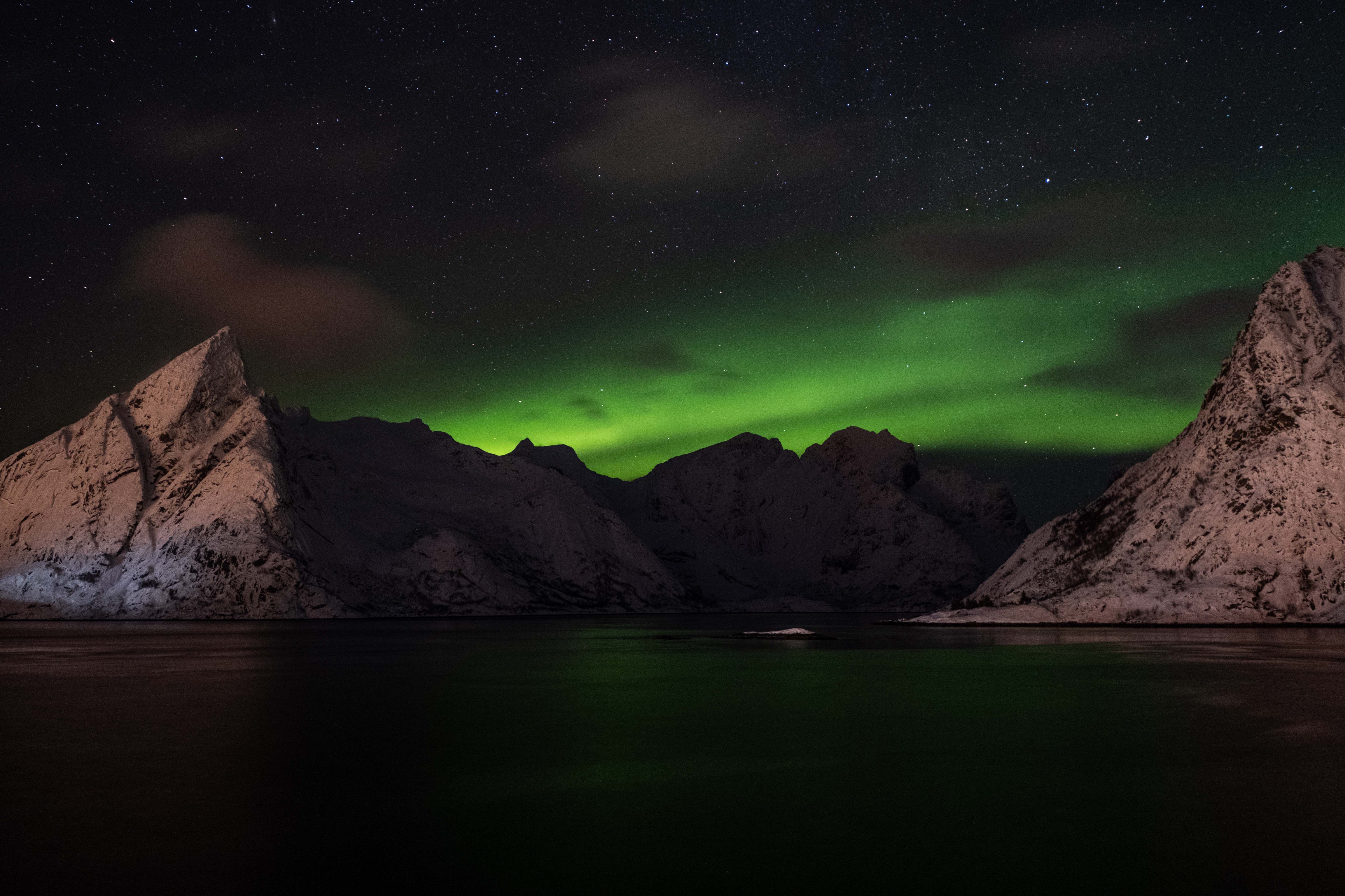 Spectacular aurora borealis illuminates the night sky over rugged snow-capped mountains, reflecting on a calm sea.