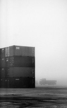 Black and white photo of containers and truck in foggy industrial area.