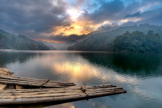 Peaceful sunrise over a tranquil lake with a bamboo raft, surrounded by lush greenery and hills.