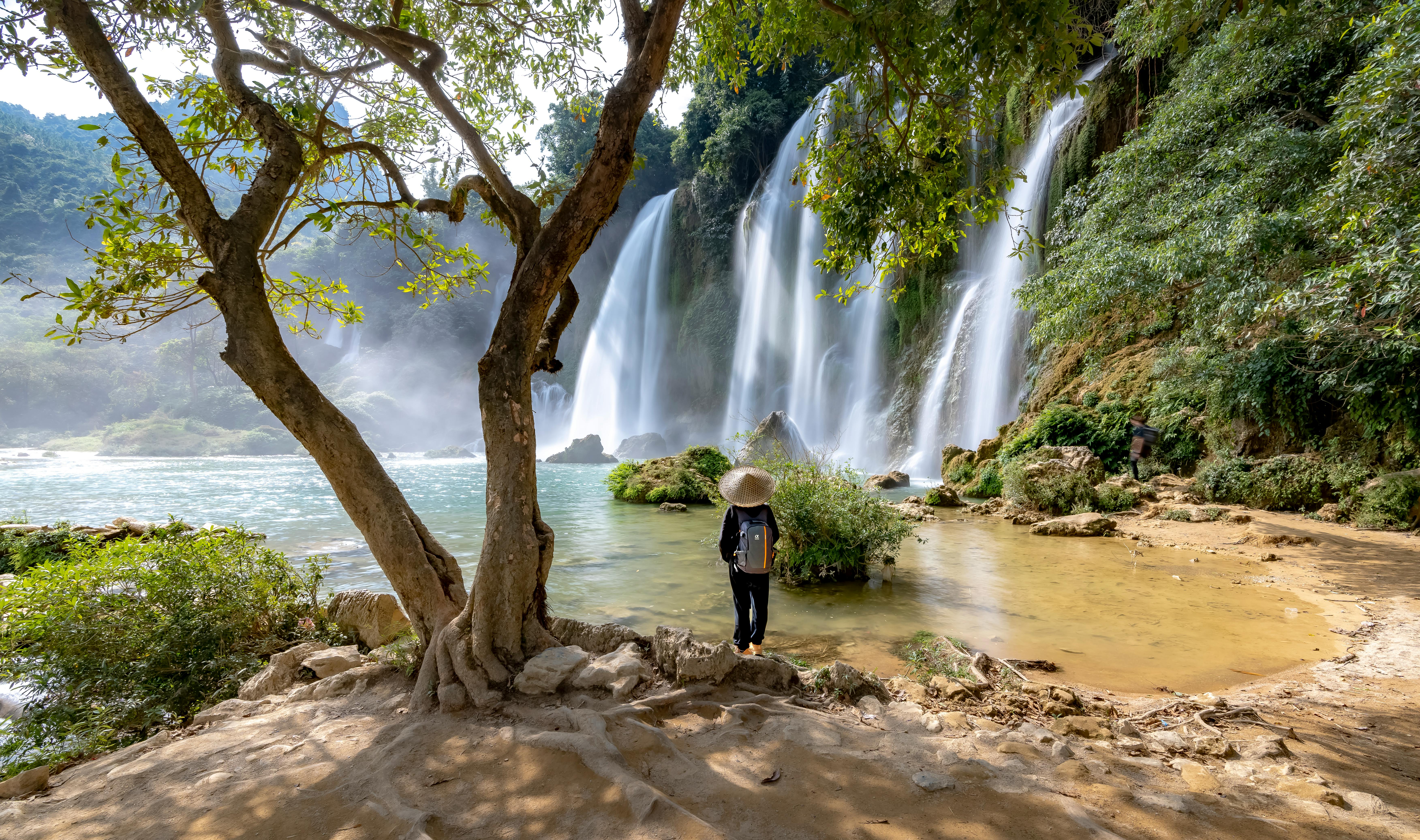 A lone traveler stands by the majestic Ban Gioc waterfall, surrounded by lush greenery.