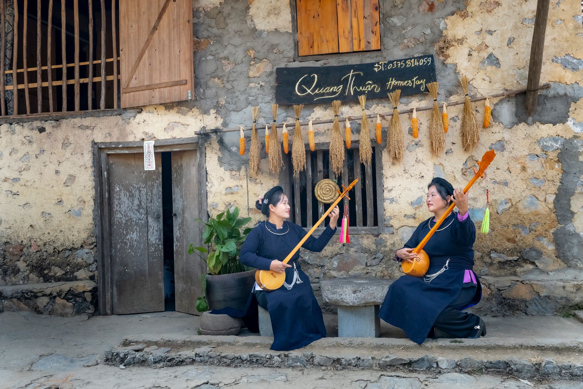 Women in traditional attire playing guitars in a rustic Vietnamese setting outdoors.
