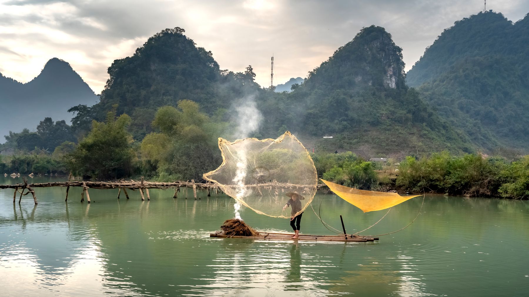 A fisherman skillfully casts a net on a tranquil river surrounded by lush mountains in Vietnam.