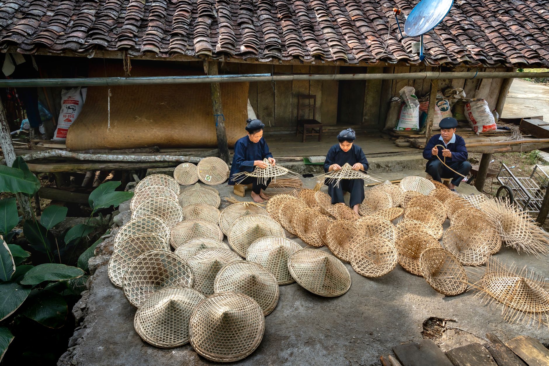 Three artisans weaving bamboo baskets in a rustic outdoor setting.