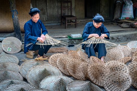Two women skillfully weave bamboo baskets in a traditional rural setting.