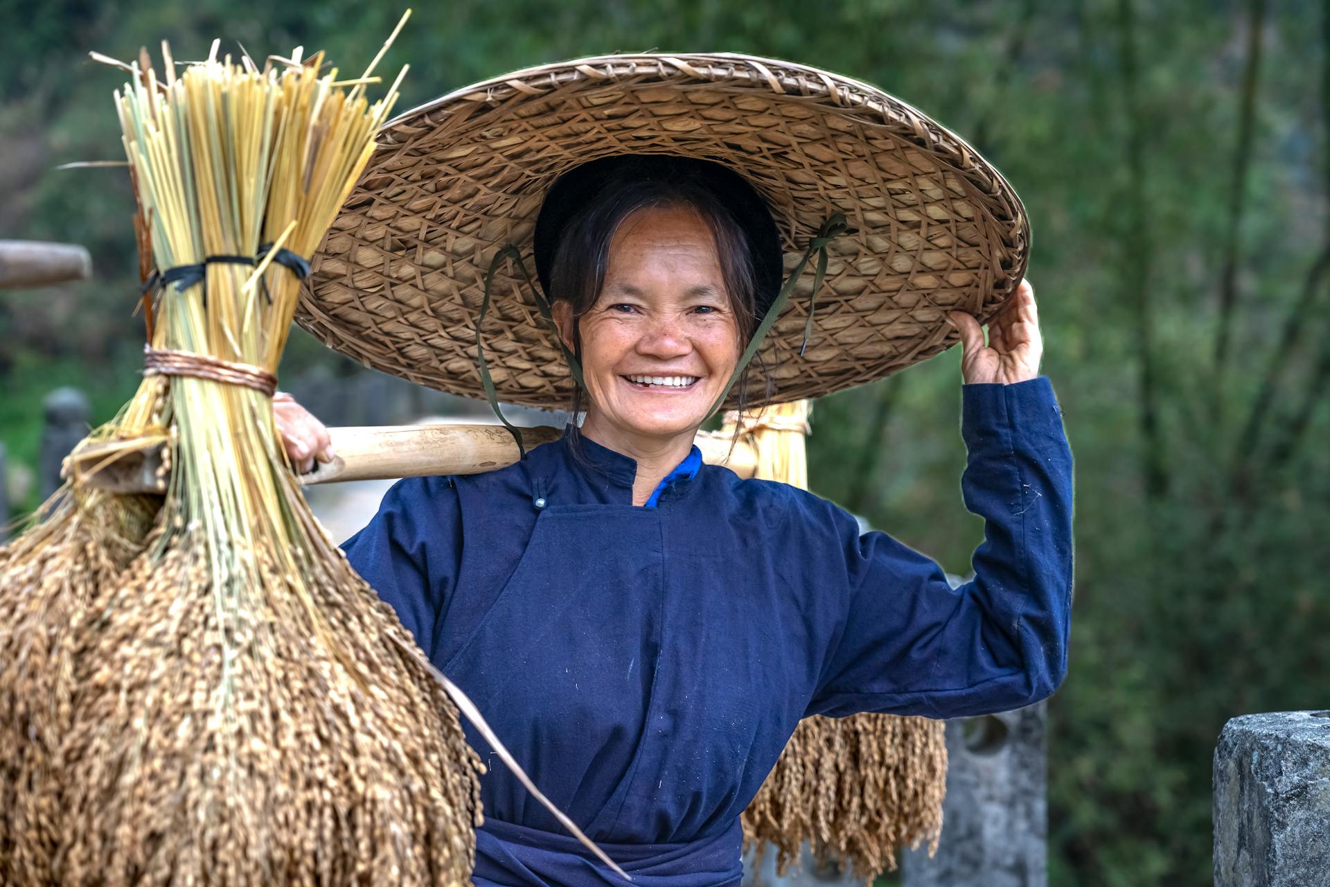 Cheerful farmer carrying rice bundles wearing a traditional straw hat outdoors.